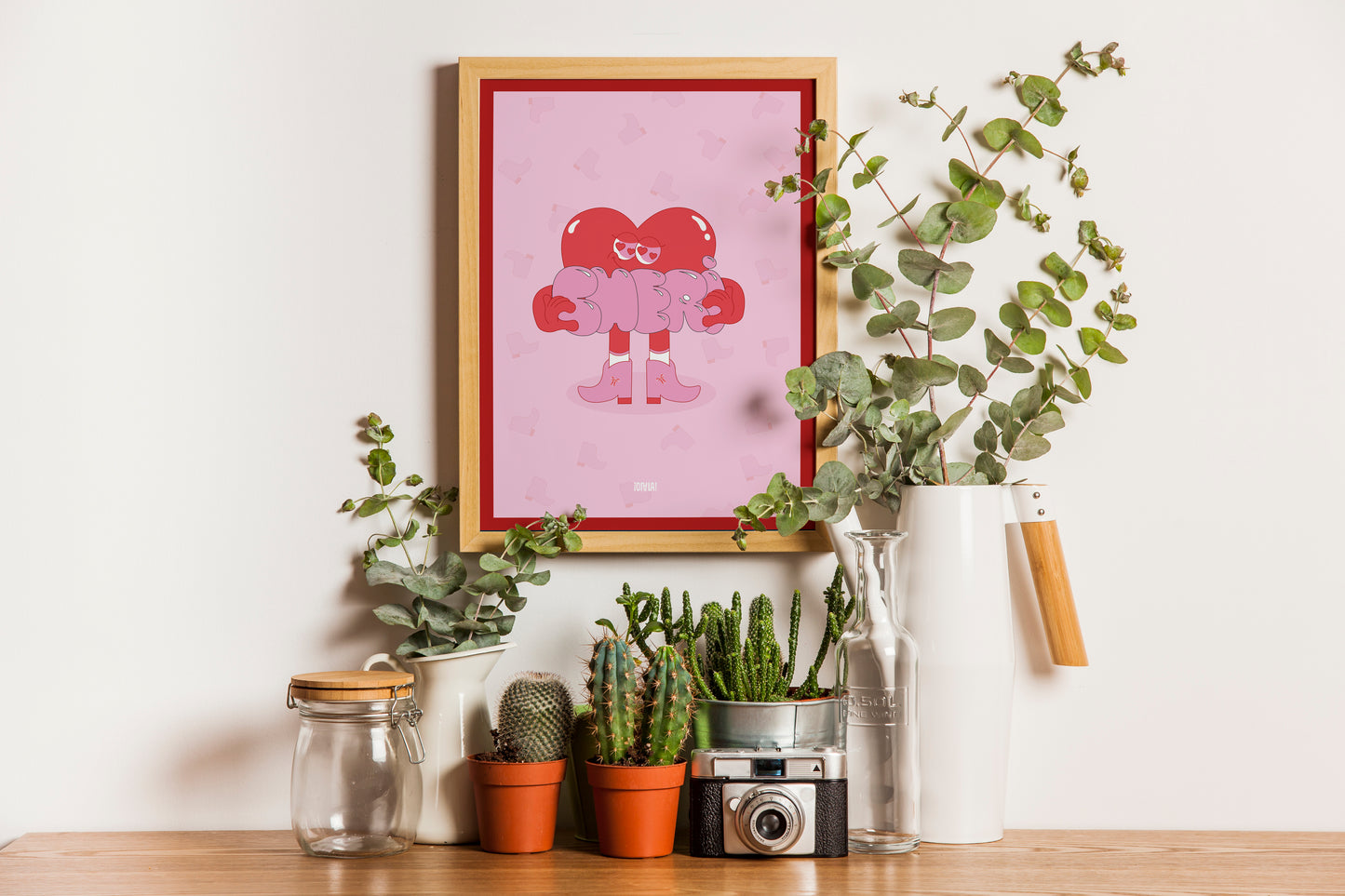 Photo de l'affiche chéri dans un cadre avec des plantes en dessous. L'affiche a un fond rose avec des santiags. Au centre se trouve un bonhomme coeur rouge qui porte dans santiags roses et tient le mot chéri dans ses mains.