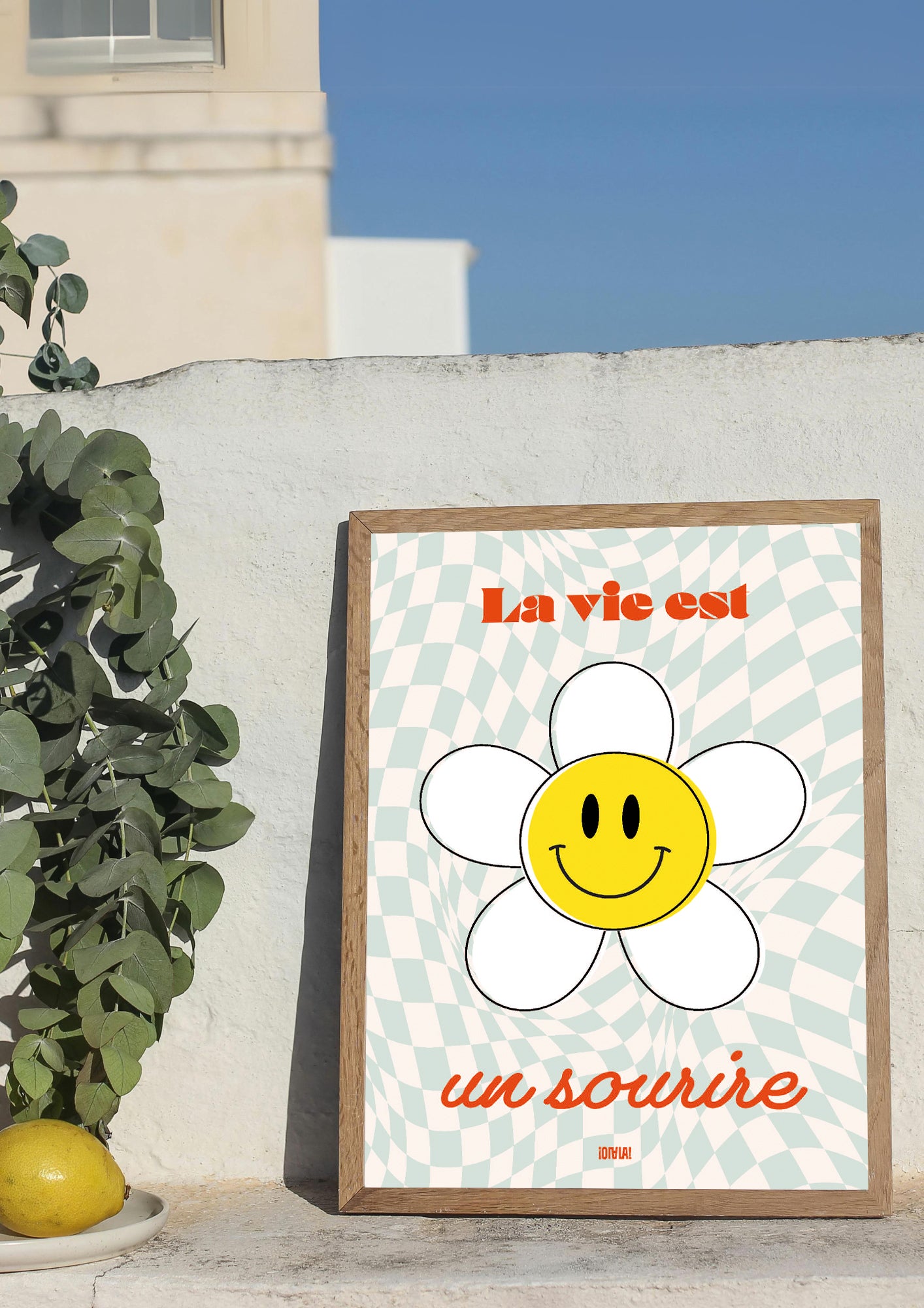 Photo produit d'une affiche dans un cadre. Le design de l'affiche est un fond quadrillé bleu et blanc cassé avec la phrase la vie est un sourire en rouge et une fleur blanche avec au centre un smiley jaune.